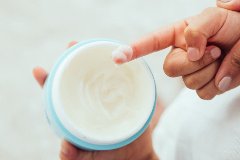 Person holding a jar of white cream with a baby's hand reaching towards it.